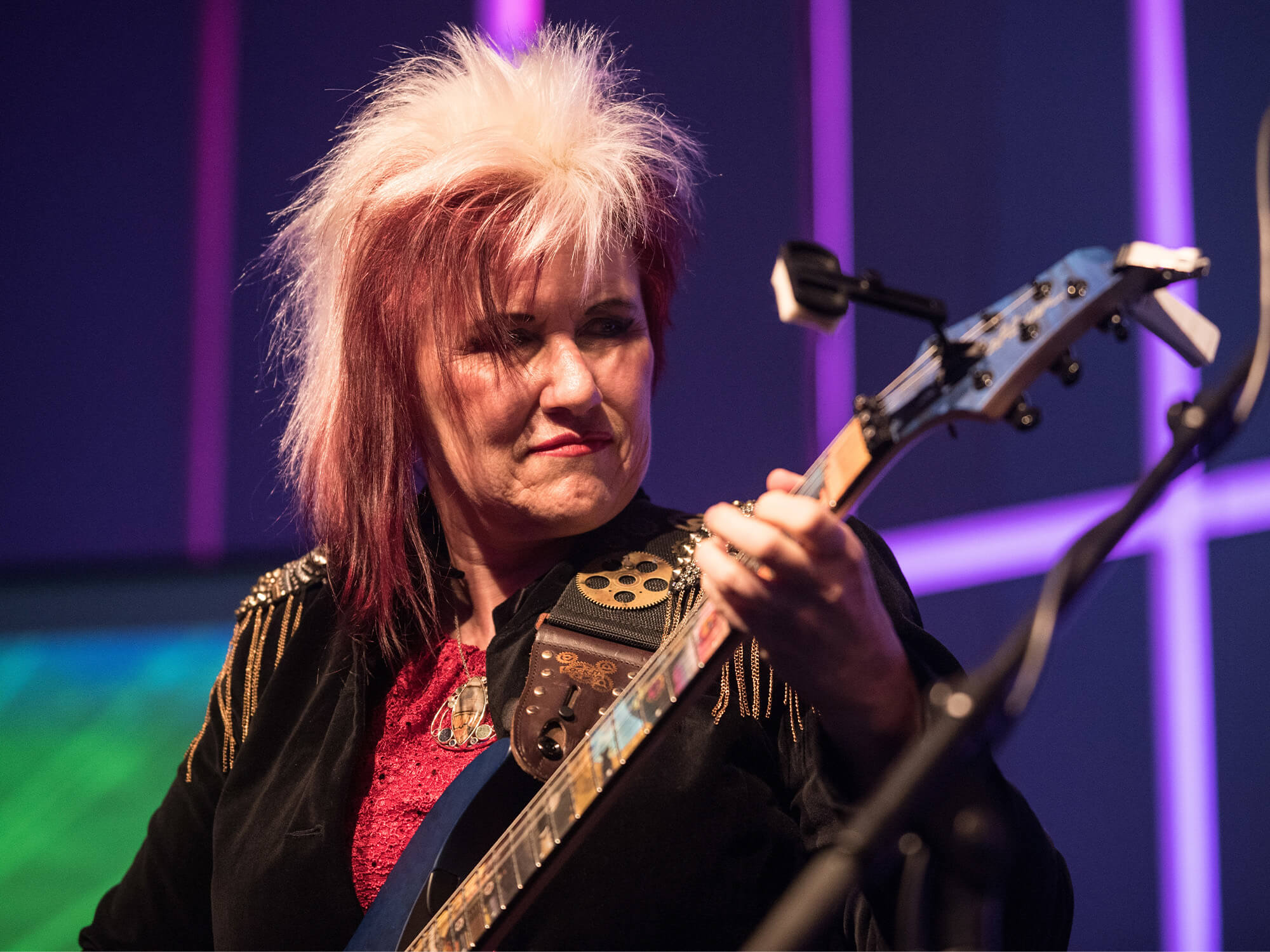 Jennifer Batten on stage. She is looking directly at her fretboard as she is playing and has vibrant pink in her hair.